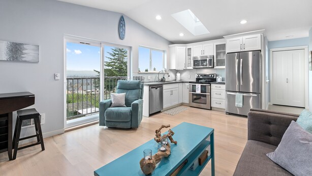 A modern kitchen with stainless steel appliances, a dining area with a blue table, and a balcony with a view. - Coastal View Home - Newly Remodeled.  Water Taxi Ride Away to see the WORLD CUP! (Seattle)