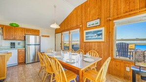 A wooden interior with a dining table set for four, kitchen cabinets, a refrigerator, and large windows offering a view. - Oceanfront Bella Beach Box - Dog Friendly w/ Hot Tub and Direct Beach Access (Avon)