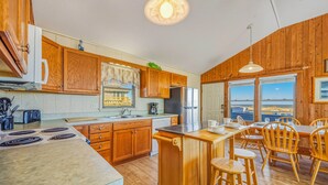 A kitchen with wooden cabinets, a stainless steel refrigerator, a coffee maker, and a view of the beach through large windows. - Oceanfront Bella Beach Box - Dog Friendly w/ Hot Tub and Direct Beach Access (Avon)