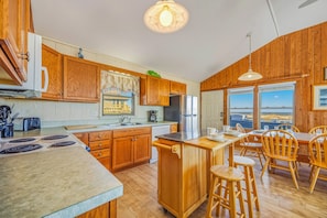A kitchen with wooden cabinets, a stainless steel refrigerator, a coffee maker, and a view of the beach through large windows. - Oceanfront Bella Beach Box - Dog Friendly w/ Hot Tub and Direct Beach Access (Avon)