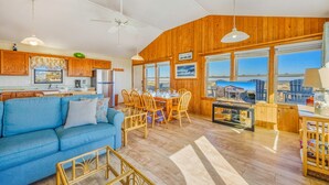 A cozy interior with a blue sofa, wooden dining table, and chairs, a kitchen area, and large windows offering a view. - Oceanfront Bella Beach Box - Dog Friendly w/ Hot Tub and Direct Beach Access (Avon)