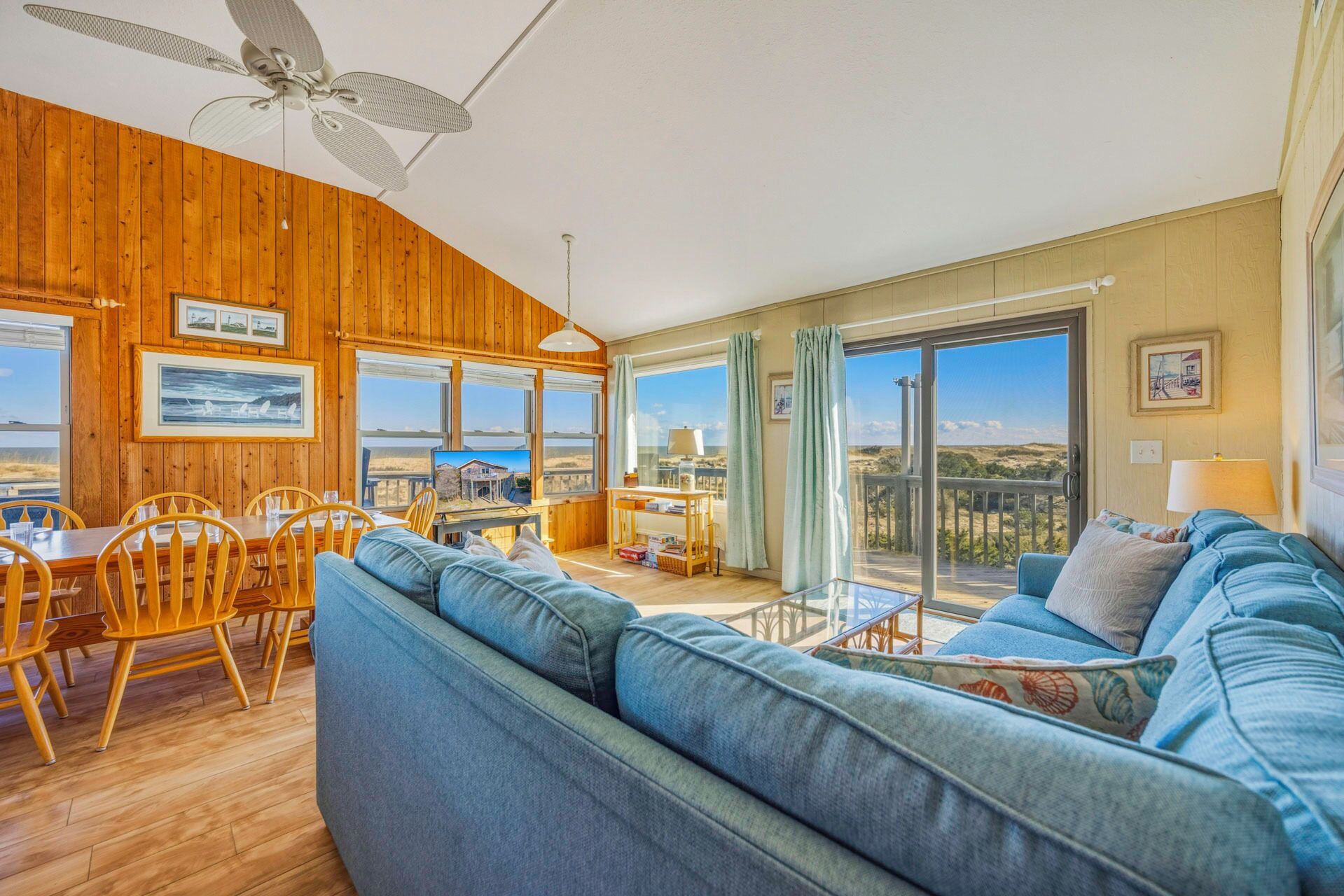 A spacious living room with a large sofa, wooden walls, a dining area, and a balcony with a view.