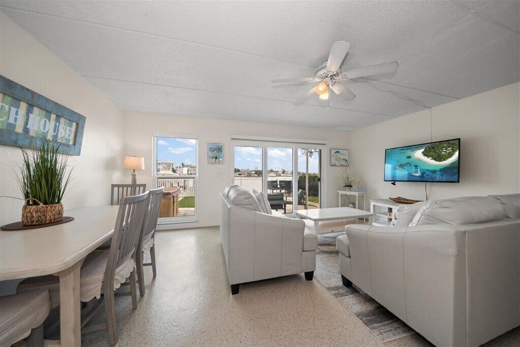 A spacious living room with a white sofa, a dining table with chairs, a ceiling fan, and a TV mounted on the wall.
