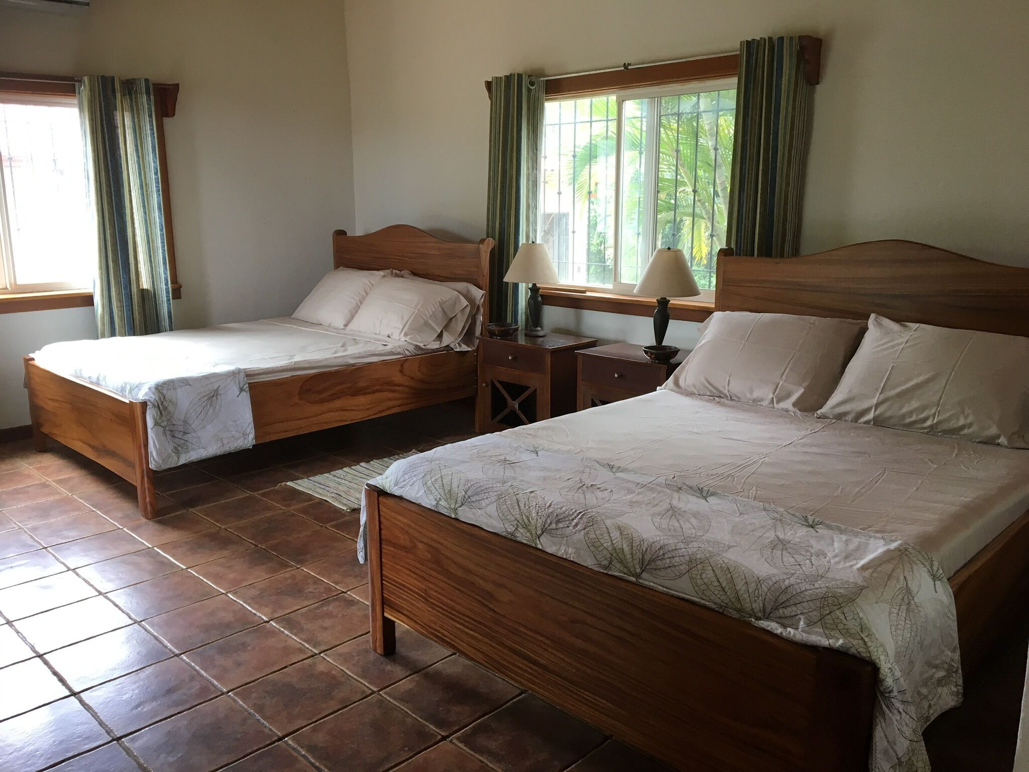 Une chambre avec deux lits en bois, chacun recouvert d’une courtepointe blanche et agrémenté d’oreillers, une table de chevet en bois avec une lampe, et une fenêtre avec des rideaux.