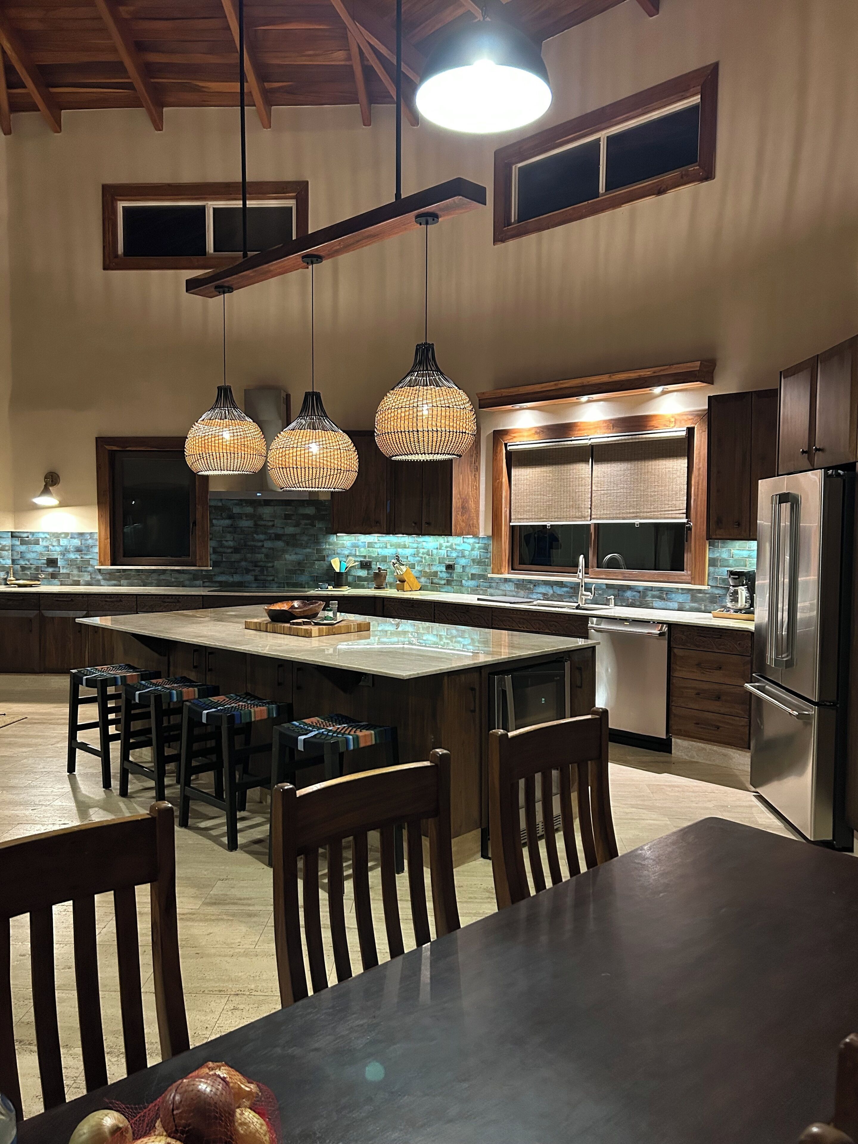 A spacious kitchen with a large island, wooden chairs, hanging pendant lights, and a stainless steel refrigerator.