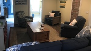 Un salon comprenant un canapé, des fauteuils, une table basse et un téléviseur. On aperçoit également un coin cuisine avec des placards et un plan de travail, le tout visible à travers une porte ouverte.