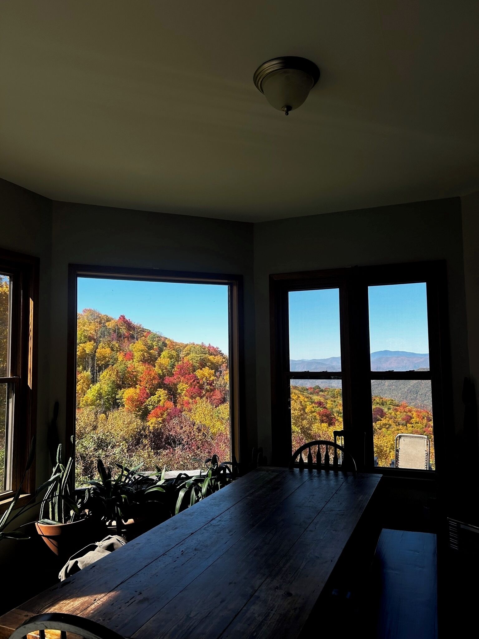 A room with a large wooden table and two windows offering a view of autumn foliage and mountains.