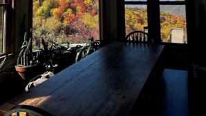 A room with a large wooden table and two windows offering a view of autumn foliage and mountains.