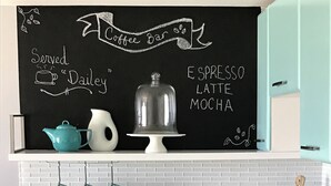 Una cocina con un menú en pizarra que enumera café y productos para el desayuno, una cafetera y una cesta de pan.