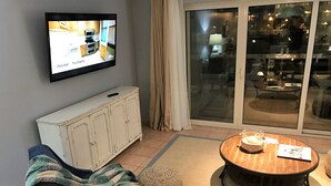 Un salón con un televisor instalado en la pared mostrando el interior de una casa. Hay un sofá, una mesa redonda de madera y un armario.
