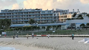 Un hotel frente a la playa con varios pisos, una playa arenosa en primer plano y cielo despejado.