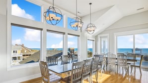 A spacious dining area with a large table and multiple chairs, large windows offering ocean views, and modern pendant lighting. - 5460- Virginia Dare, 8BR Oceanfront Retreat | Pool, Theater & Game Room (Nags Head)
