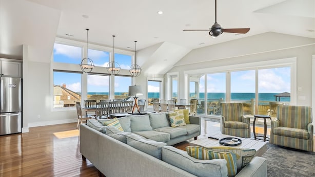 A spacious living room with a large gray sectional sofa, a dining area with chairs, and a view of the ocean through large windows. - 5460- Virginia Dare, 8BR Oceanfront Retreat | Pool, Theater & Game Room (Nags Head)