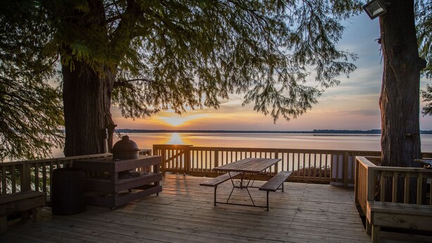 A wooden deck with picnic tables and benches overlooking a calm body of water during sunset.