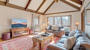 A spacious living room with a high ceiling, wooden beams, a large TV, comfortable seating, and large windows.