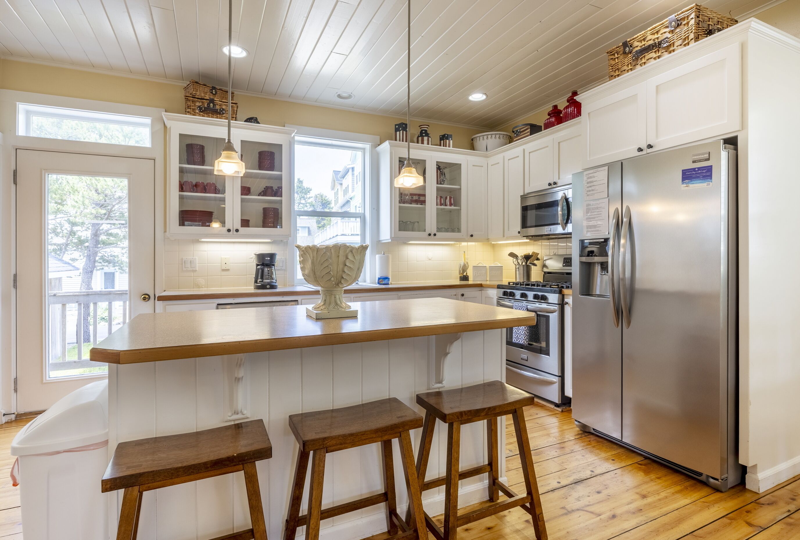Una cocina con gabinetes blancos, una isla central con taburetes, un refrigerador de acero inoxidable y una ventana con vistas al exterior.