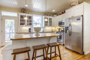 A kitchen with white cabinets, a central island with stools, a stainless steel refrigerator, and a window with a view of the outside.