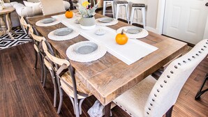 A dining area with a wooden table set for four, surrounded by upholstered chairs. A chandelier hangs above, and a kitchen is visible in the background. - Modern Farmhouse Just Steps From Town and Lake Michigan w/Heated Pool! (New Buffalo)