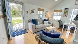 A living room with a grey sofa, blue cushions, a wicker chair, a small table, and a door leading to a garden.