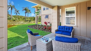 A covered patio with wicker furniture, a glass table, and a view of a well-maintained lawn and palm trees. - Pili Mai at Poipu #03D By Parrish Kauai - Great Resort pool for families (Koloa)