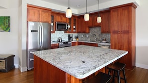A kitchen with wooden cabinets, a stainless steel refrigerator, a microwave, a stove, and a marble island with bar stools. - Pili Mai at Poipu #03D By Parrish Kauai - Great Resort pool for families (Koloa)