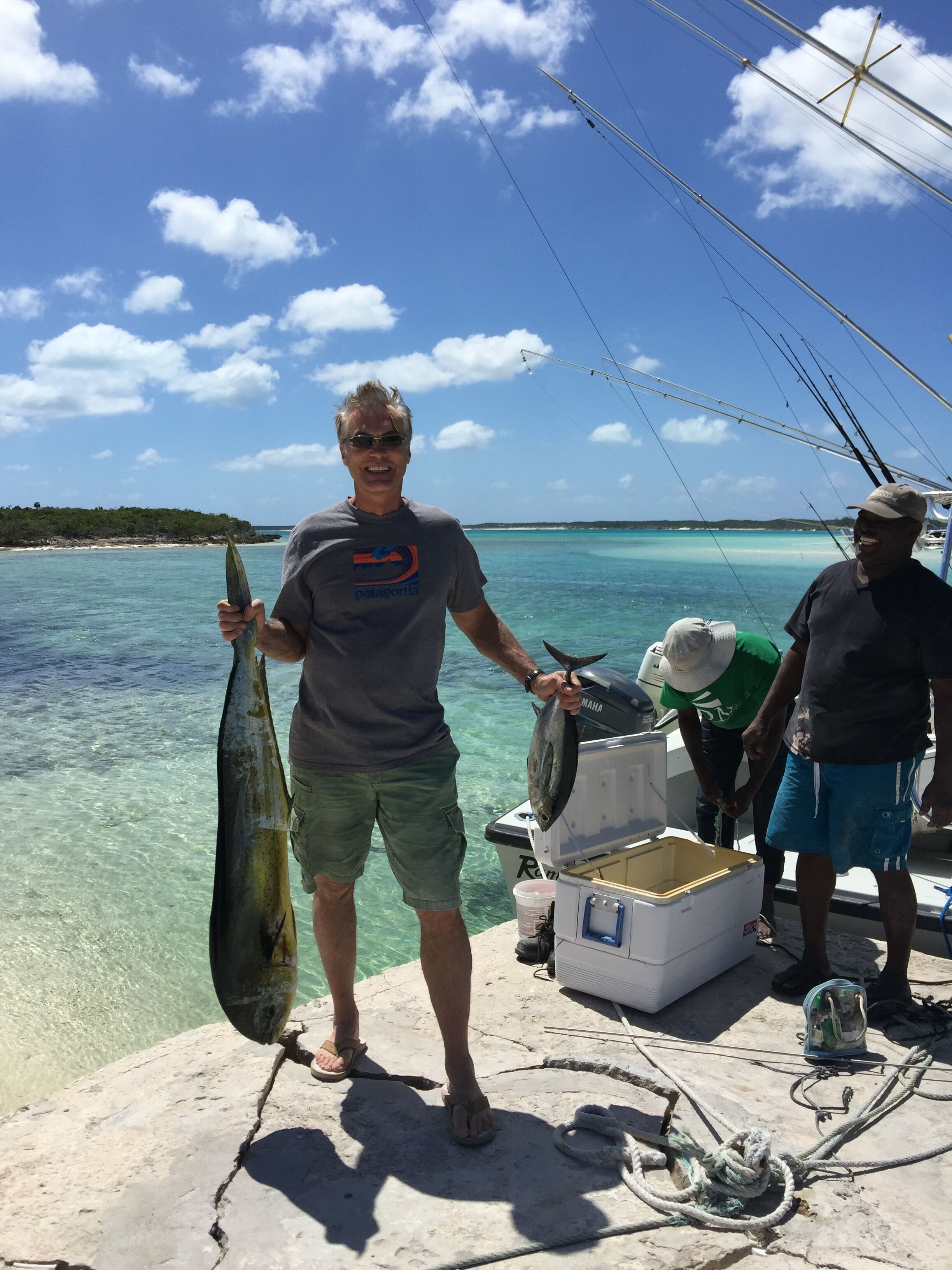 En person håller i en fisk på en båt, med klart blått vatten och en stenig strand i bakgrunden.