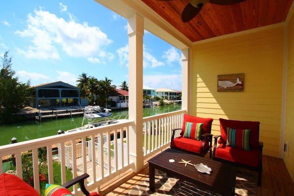 A covered outdoor area with a wooden deck, a red sofa with colorful cushions, and a view of a marina with boats and houses in the background.