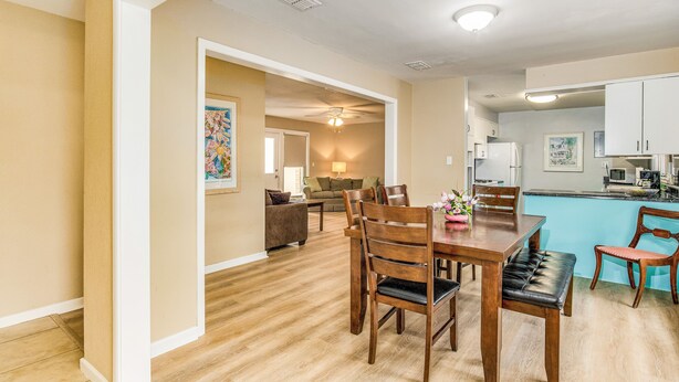 A dining area with a wooden table and chairs, a kitchen with white cabinets, and a living room with a sofa and a ceiling fan. - Near EVERYTHING House-Now Discounted for longer stays (Pensacola)