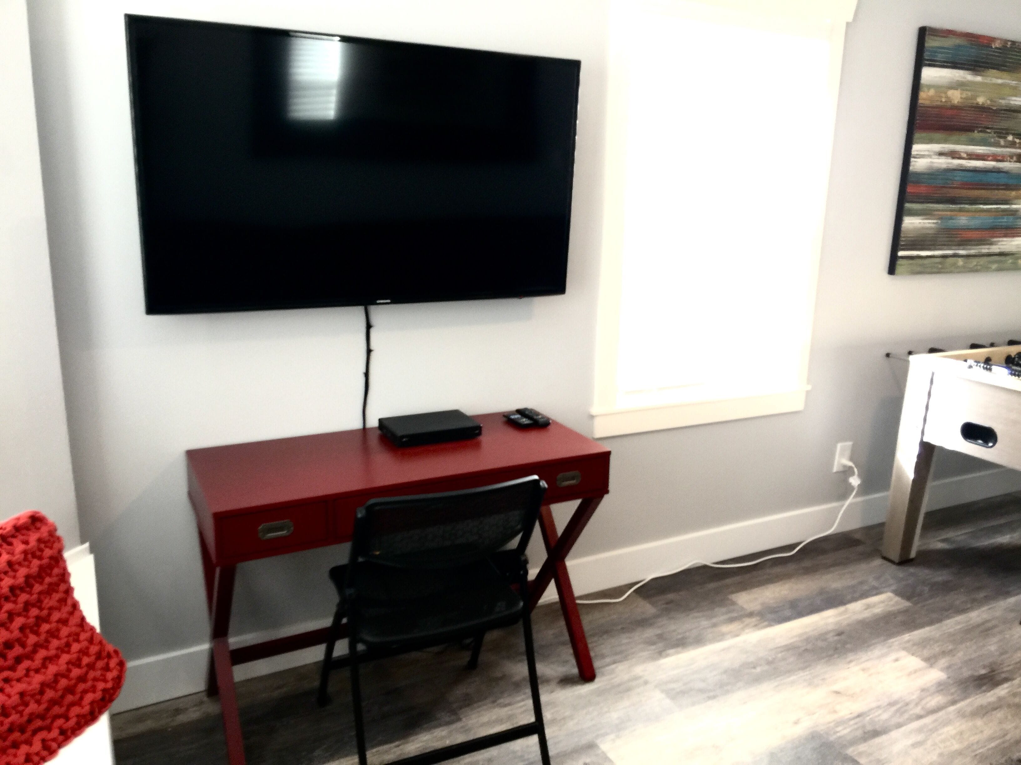 Una habitación con un escritorio rojo, una silla negra, un televisor de pantalla plana montado en la pared y una mesa de fútbol.