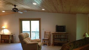 A living room with a recliner, a television on a wooden stand, a ceiling fan, and a window with a view of greenery.