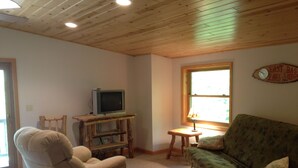 A cozy living room with a wooden ceiling, a television on a wooden stand, a green sofa, and a recliner.