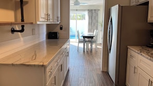 A modern kitchen with white cabinetry, a marble countertop, and a view to a dining area with a table and chairs. - Anna Maria 2 Family Home w/ pool, beach/3 blocks. (Holmes Beach)