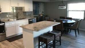 Une cuisine moderne avec des meubles blancs, un plan de travail en granit et un coin repas agrémenté de chaises en osier et d’un lustre.