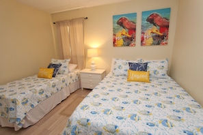 Quarto com duas camas de solteiro, cada uma com um edredom estampado e almofadas a combinar, uma mesa de cabeceira com abajur e uma janela com cortina.