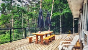 Eine Holzterrasse mit Tisch und Bänken, umgeben von einem schwarzen Geländer, mit Blick auf einen bewaldeten Bereich.