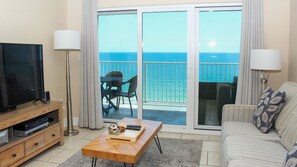 A living room with a TV, a coffee table, a sofa, a balcony with a view of the ocean, and a dining area with chairs.