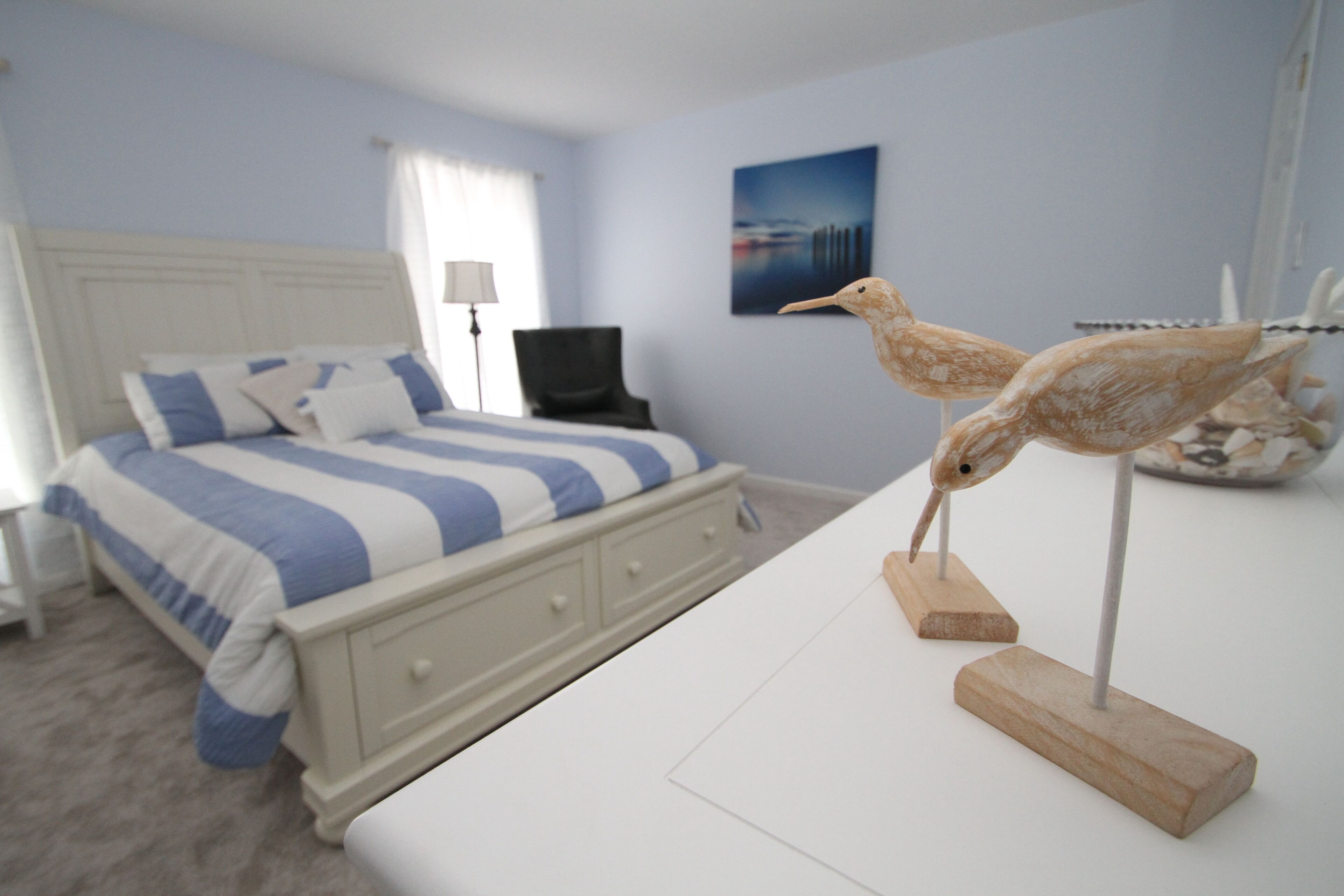 Een slaapkamer met een bed, een dressoir, een lamp en twee houten beeldjes van vogels.