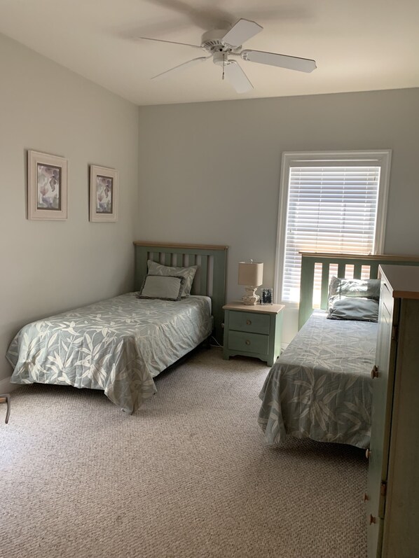 A bedroom with two single beds, a nightstand with a lamp, a ceiling fan, and two framed pictures on the wall.