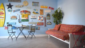 Ein Zimmer mit blauer Wand, verziert mit verschiedenen Strand-Dekorationen und einem Surfbrett. Es gibt ein weißes Sofa und zwei Klappstühle.