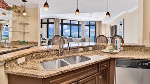 A modern kitchen with granite countertops, stainless steel sinks, and a dishwasher. There are pendant lights, a large window with a view, and a seating area with a view. - The Oasis 2010 | Spacious, Lazy River, Poolside Grill, New Splash Pad, Pool with Slide! (Orange Beach)