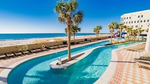 A swimming pool with a curved design, palm trees, and a beach view. - The Oasis 2010 | Spacious, Lazy River, Poolside Grill, New Splash Pad, Pool with Slide! (Orange Beach)