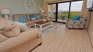 A spacious living area with a sofa set, a coffee table, and a patterned armchair. A large window offers a view of the outdoors.