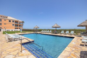 A swimming pool with lounge chairs and umbrellas, a building with balconies, and a clear sky.