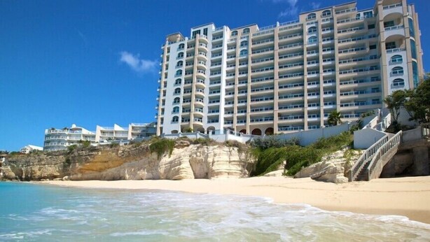 Un hotel frente a la playa con varios balcones, una playa de arena y un acantilado rocoso.