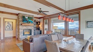A spacious living room with a stone fireplace, a large leather sofa, a dining table with chairs, and a view of the mountains through large windows.
