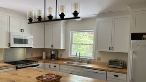 A modern kitchen with white cabinets, a wooden countertop, a built-in microwave, a coffee maker, a toaster, and a wooden island with a basket. - Tall pines & lake view. Perfect location ctr of MDI! Close trails.  Local owners (Mt Desert)