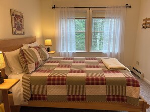 Een slaapkamer met een groot bed, een nachtkastje met lamp, een raam met dunne gordijnen en een wanddecoratie.