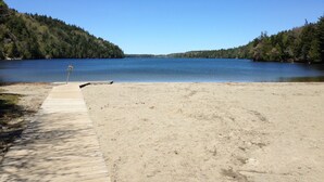A sandy beach with a wooden walkway leading to a dock, surrounded by forested hills and a calm lake. - Tall pines & lake view. Perfect location ctr of MDI! Close trails.  Local owners (Mt Desert)