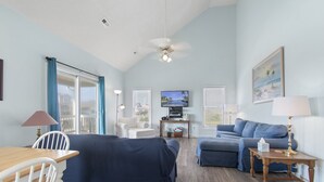 Uma sala de estar com uma mesa de madeira, cadeiras brancas, um sofá azul, uma poltrona azul, uma televisão e um ventilador de teto.