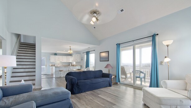 Uma sala de estar ampla, com um sofá azul escuro, uma poltrona branca e um sofá branco. Uma escada leva a uma área de cozinha ao fundo.